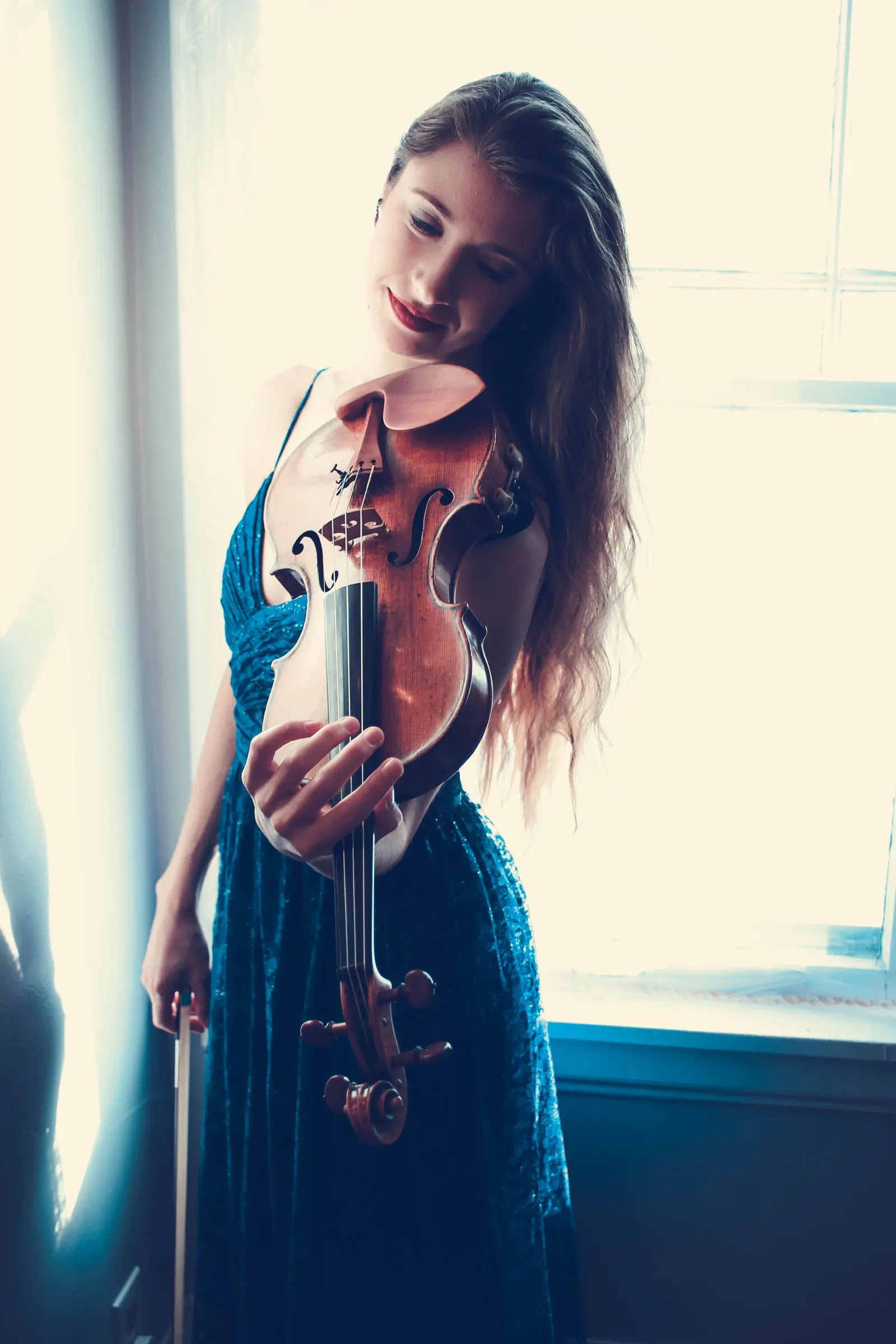 Madalyn in a teal gown by a bright window, admiring her violin
